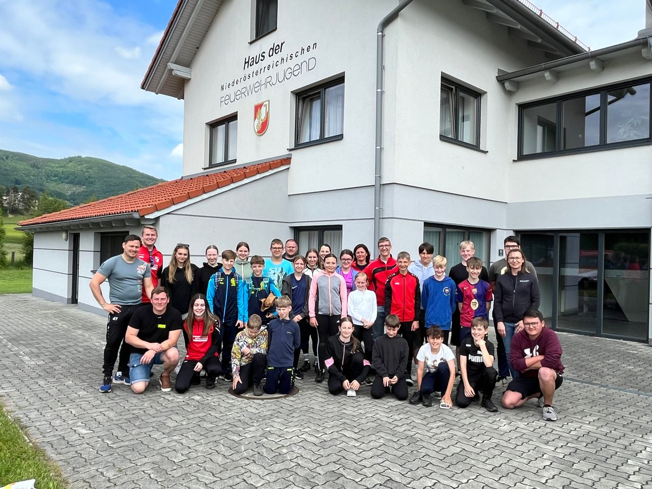 Eine Gruppe von Menschen steht vor einem Gebäude und posiert wahrscheinlich für ein Foto. Das Gebäude hat ein Schild mit der Aufschrift 'Haus der Niederosterreichischen Feuerwehrjugend.' Die Personen tragen verschiedene Jacken und einige haben Brillen. Das Gebäude hat viele Fenster und ein rotes Dach. Ein Berg ist im Hintergrund sichtbar.