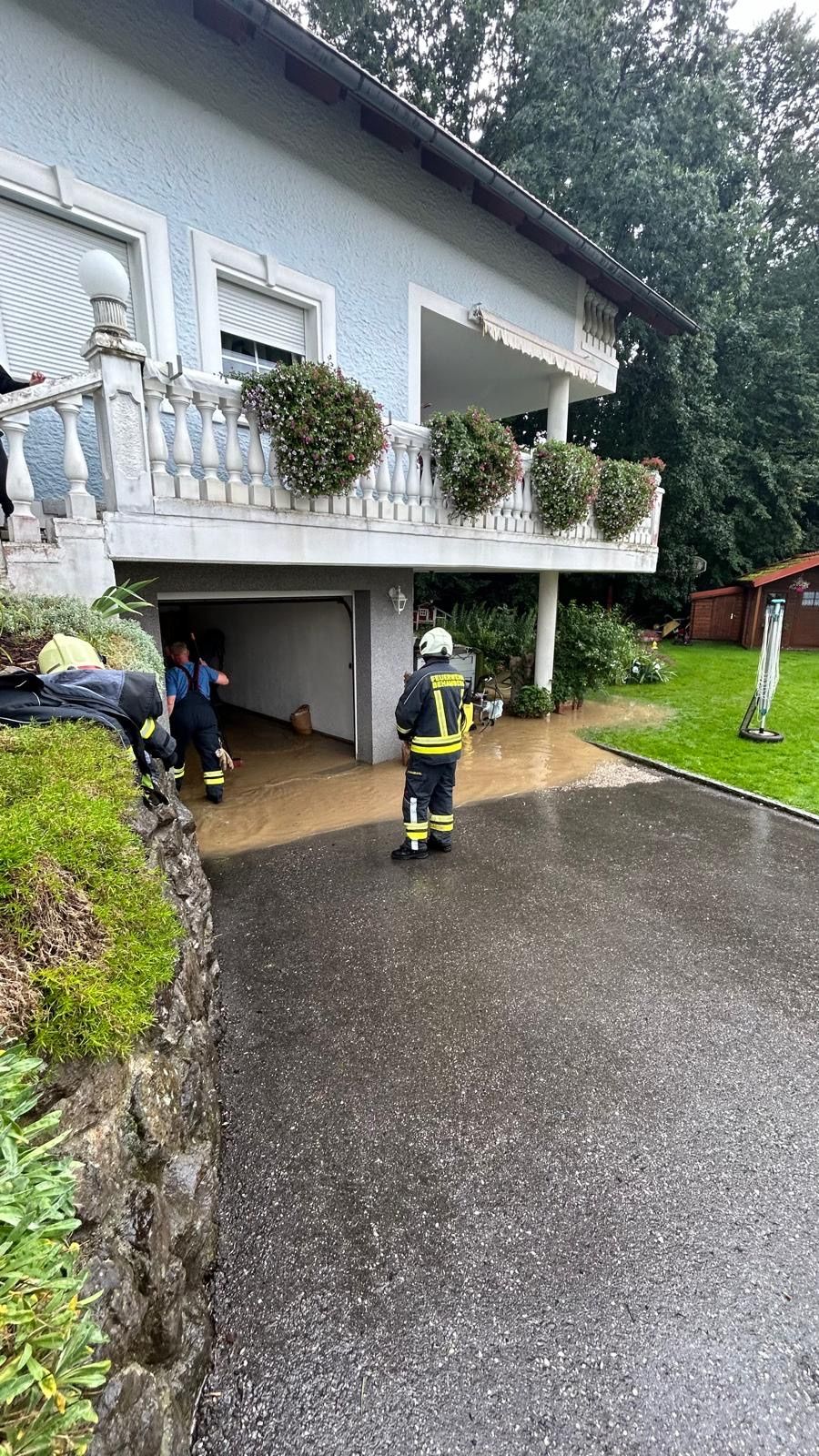 Feuerwehrleute in Uniform stehen vor einem Haus, möglicherweise nach einer Überschwemmung. Ein Feuerwehrmann hält einen Schlauch.