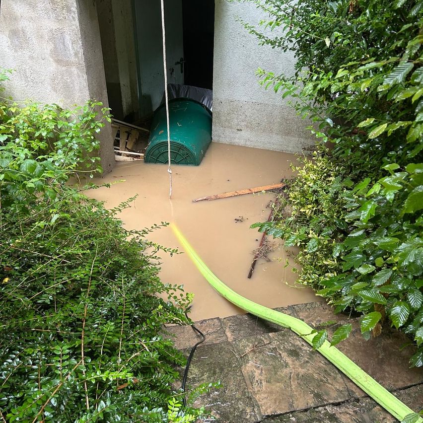 Ein überfluteter Außenbereich mit einem grünen Fass, einem Schlauch und einem Eingang, der zu einem Gebäude führt.