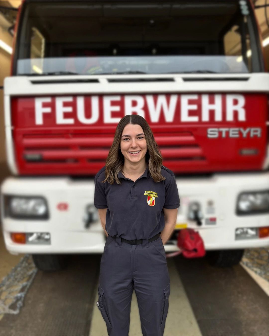 Eine Frau steht vor einem roten Feuerwehrfahrzeug mit der Aufschrift 'FEUERWEHR' auf der Vorderseite. Sie trägt eine dunkelblaue Uniform mit einem Logo auf der linken Seite.