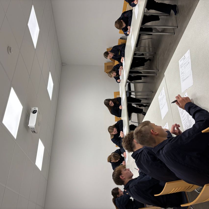 Ein Klassenzimmer mit mehreren Schülern, die an Tischen sitzen und auf Papieren schreiben. Ein Beamer ist an der Wand montiert.