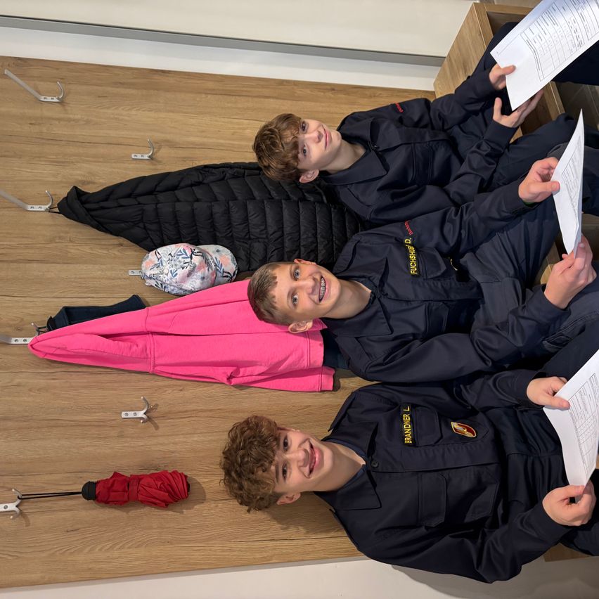 Drei Jungen in Uniformen liegen auf einem Holzboden, lächeln und halten Papiere. Sie sind von verschiedenen Gegenständen umgeben, darunter ein rosa Regenschirm und eine schwarze Jacke.