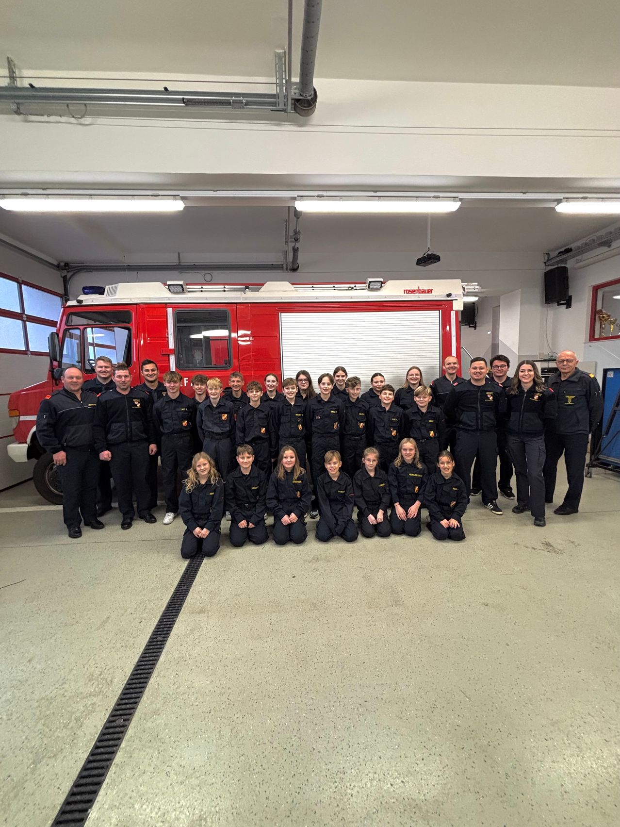 Eine Gruppe Feuerwehrleute und Kinder in Uniformen posiert für ein Foto in einer Garage mit einem Feuerwehrauto.