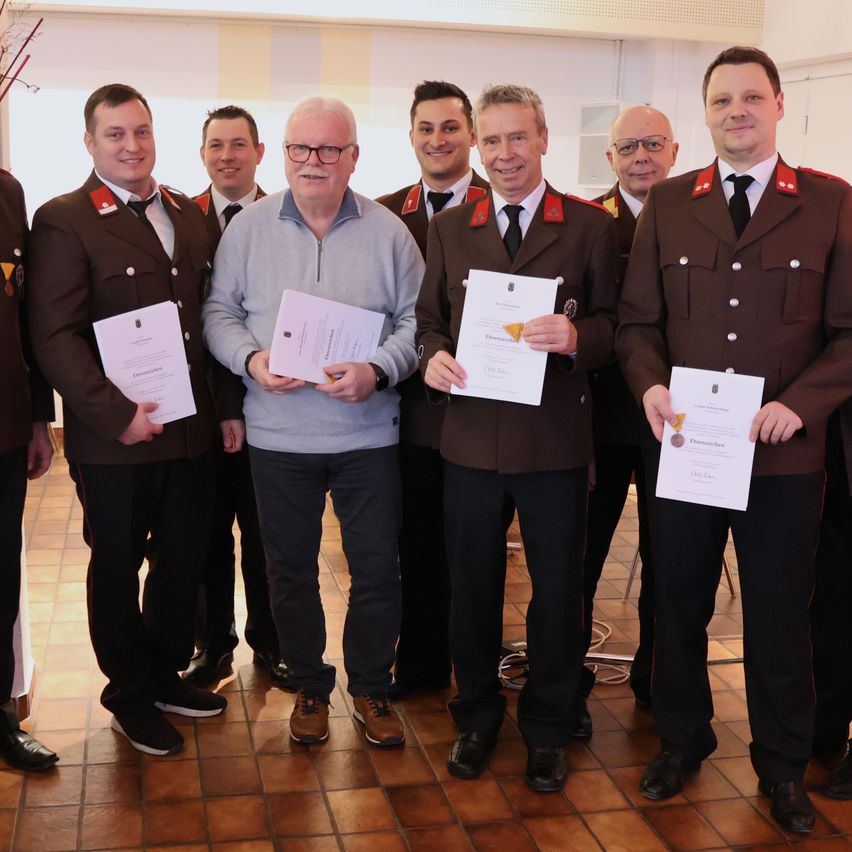 Eine Gruppe von Männern in Uniform steht zusammen und hält Urkunden in den Händen, ein Mann in einem grauen Pullover steht in der Mitte.