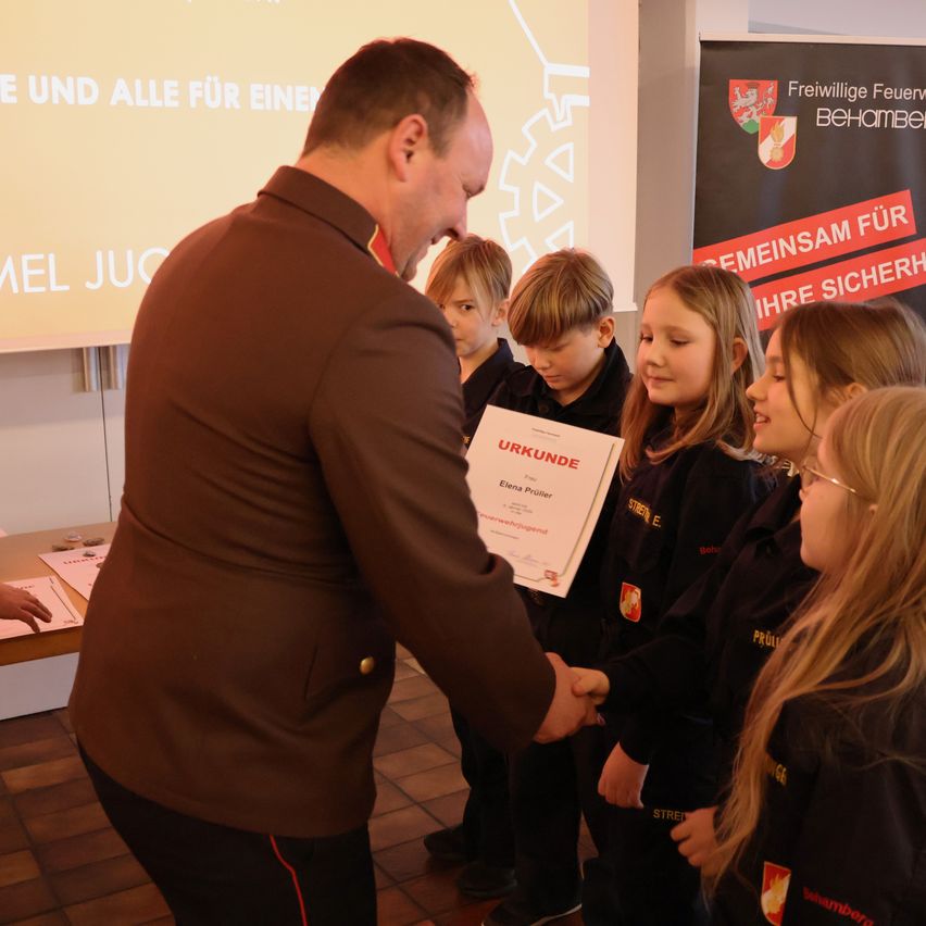 Ein Mann in Uniform überreicht einem kleinen, in schwarz gekleideten Kindern mit gelbem Text eine Urkunde. Dahinter befindet sich eine Projektionsleinwand mit deutschem Text.