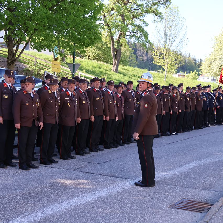 A group of uniformed individuals, likely military or police, stands in formation on a road under a clear sky. A flag is held by a person at the end of the line.