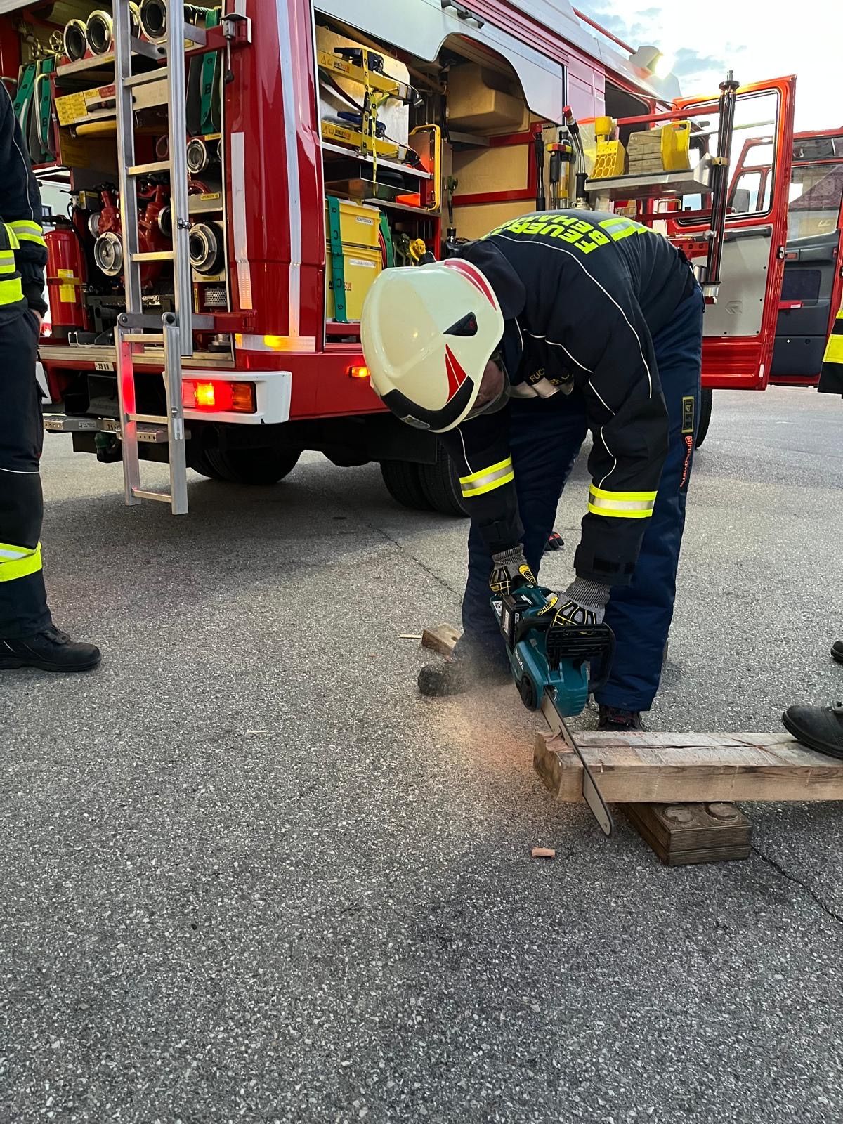 Ein Feuerwehrmann in Uniform säg mit einer Kettensäge ein Holzbrett in der Nähe eines Feuerwehrwagens.