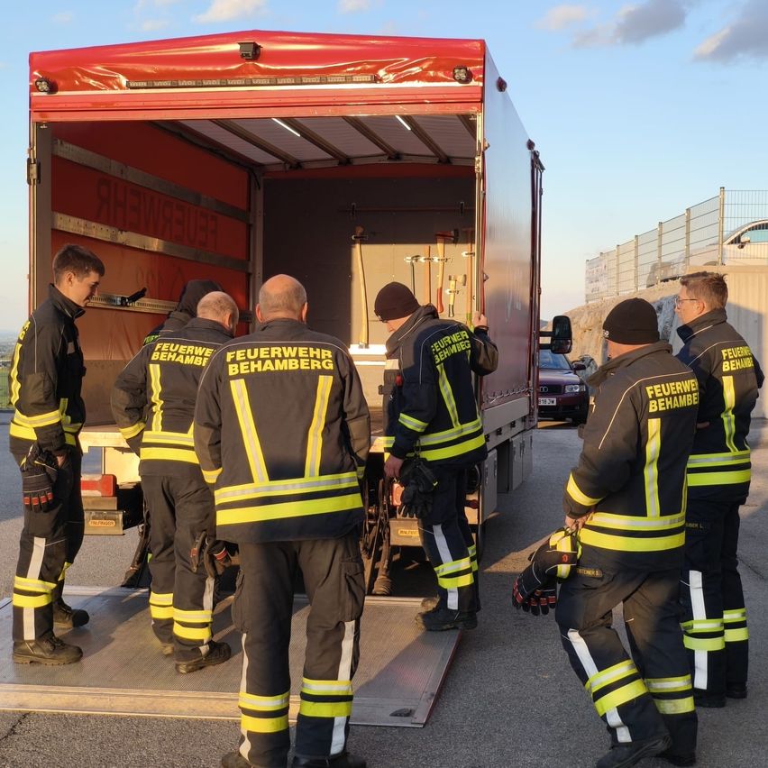 Eine Gruppe Feuerwehrleute in Uniform lädt Ausrüstung in einen Lkw. Sie stehen vor der offenen Rückseite des Trucks.