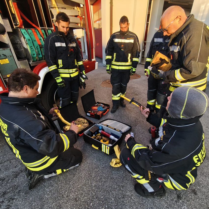 Eine Gruppe von Feuerwehrleuten ist um eine offene Werkzeugkiste versammelt und bereitet Ausrüstung vor. Sie tragen vollständige Ausrüstung, einer hat eine Mütze. Hinter ihnen steht ein Feuerwehrwagen mit Ausrüstung.