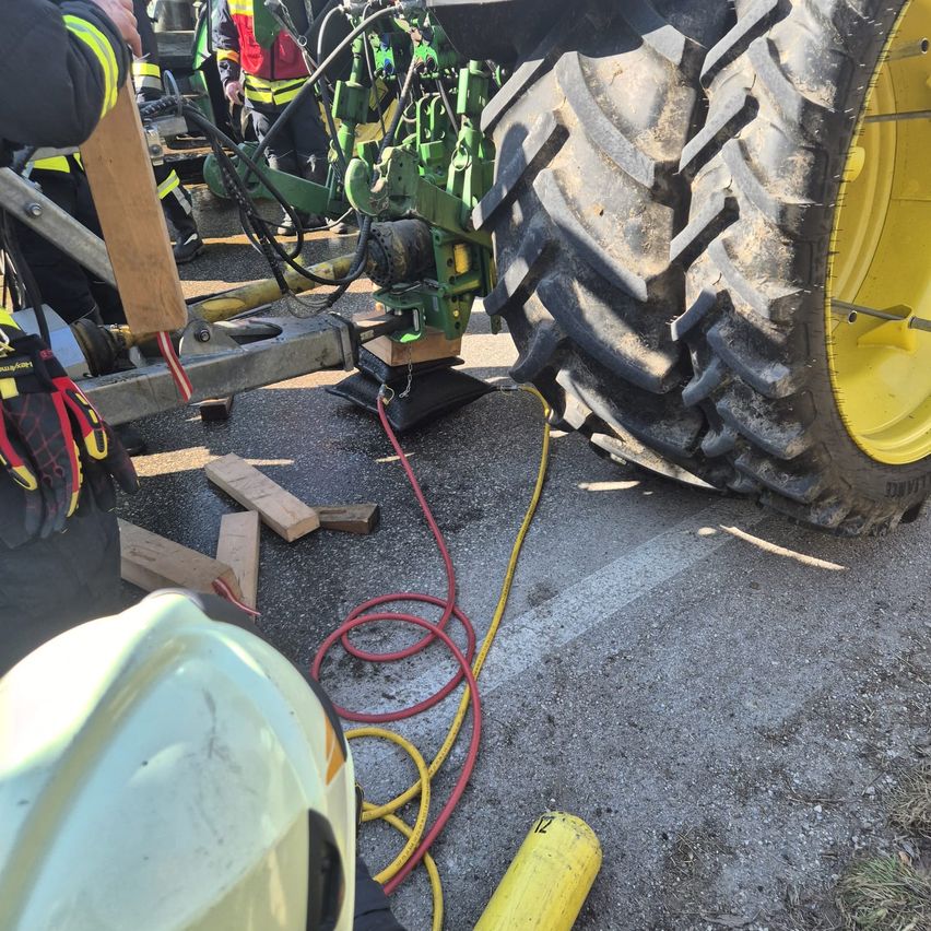 Feuerwehrleute verwenden Werkzeuge, um einen Traktor zu stabilisieren. Unter dem Rad befinden sich Holzplanken, und Kabel sind angeschlossen. Ein gelber Sicherheitshelm ist in der Nähe.