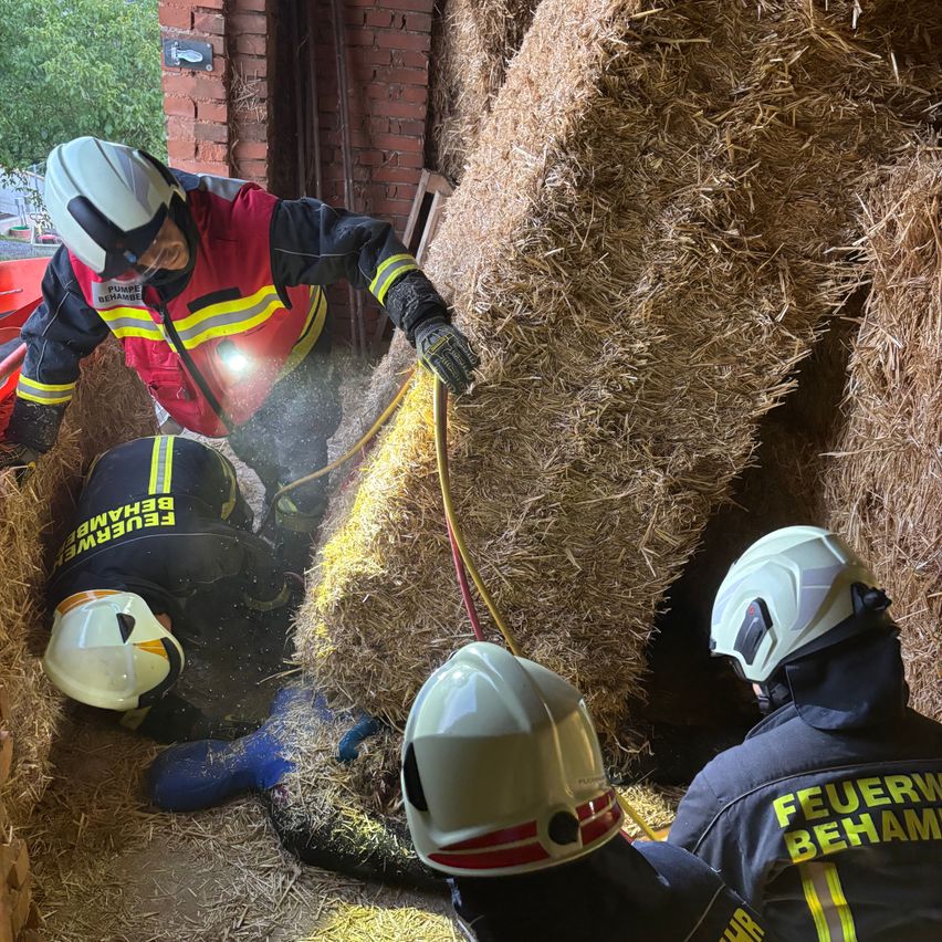 Three firefighters in protective gear are rescuing someone buried in hay bales. One is lying on the ground, another is standing, and the third is reaching out.