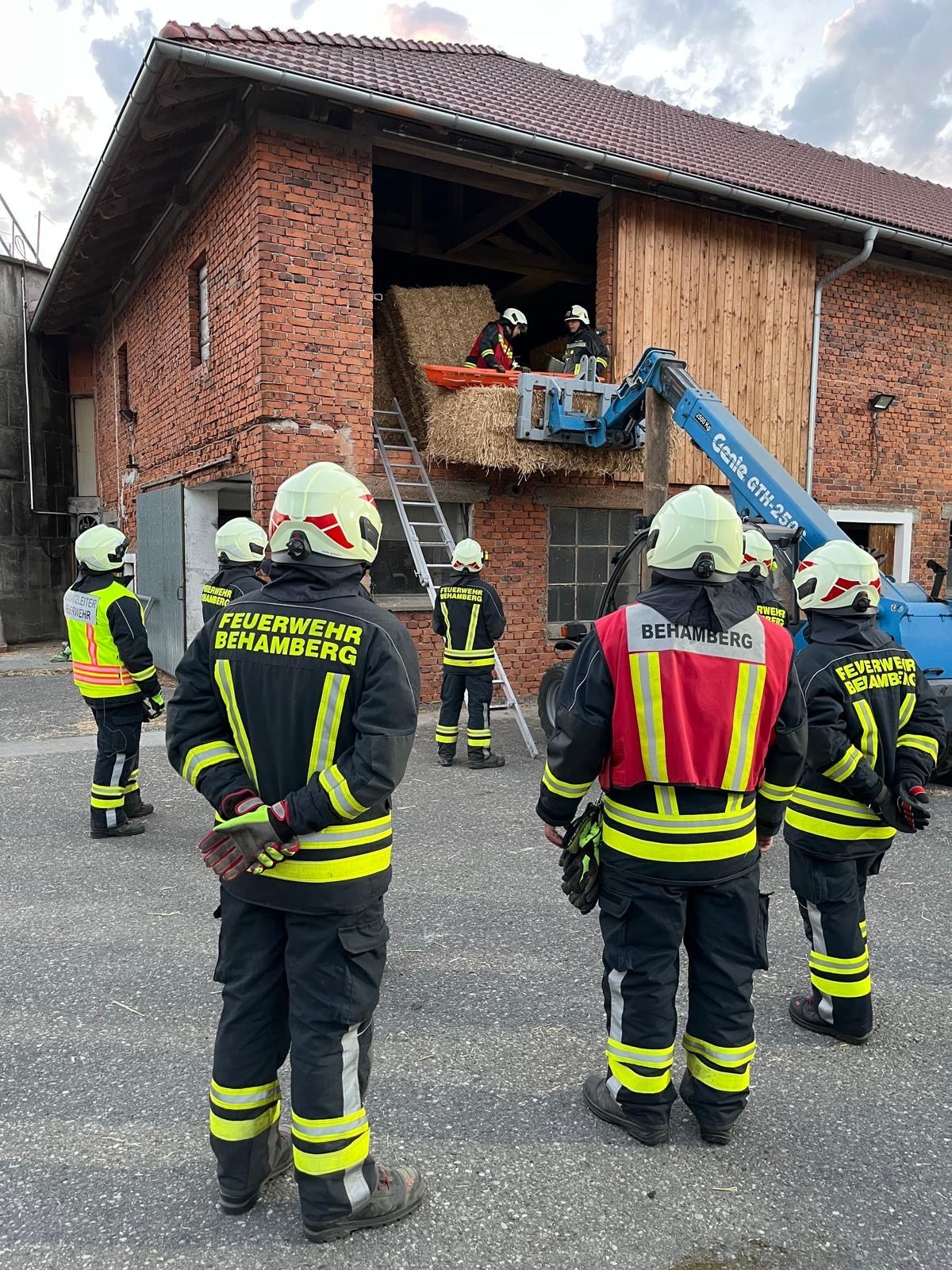Firefighters in full gear stand outside a brick building, with two individuals inside using a crane to remove a hay bale.
