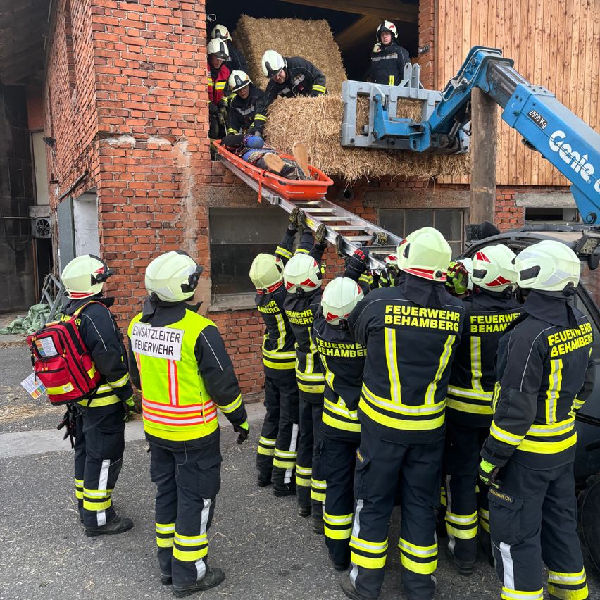 A group of firefighters in yellow and black uniforms are gathered around a building with a brick wall. One person is on a stretcher being lowered from the building by a crane.
