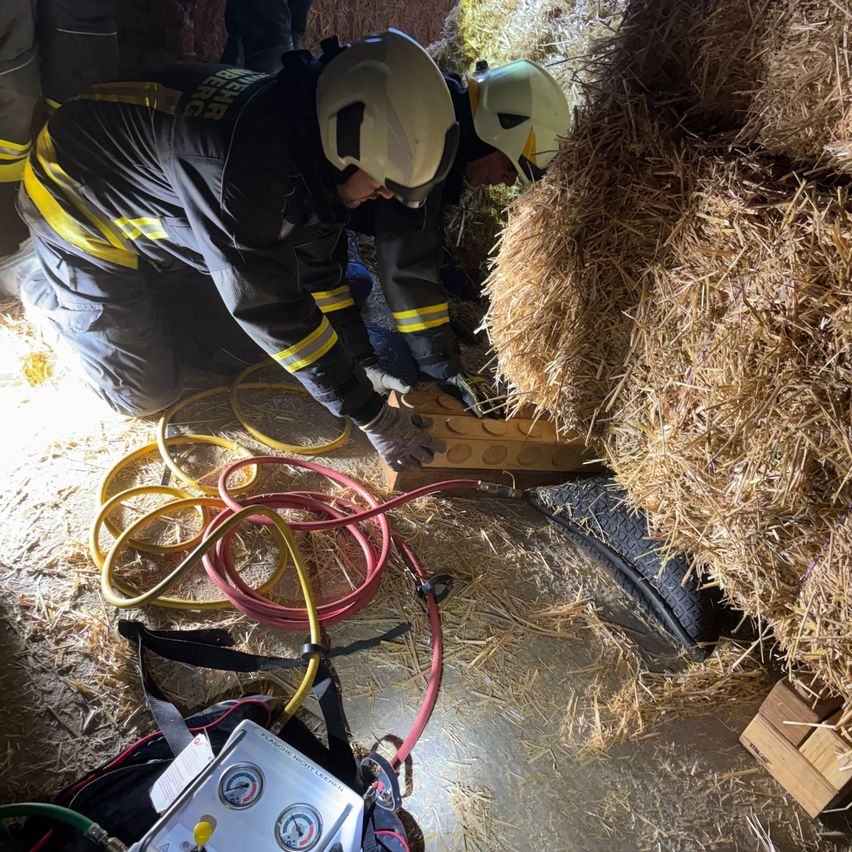 Two firefighters in protective gear are working on a rescue operation in a barn, surrounded by hay bales and various tools.