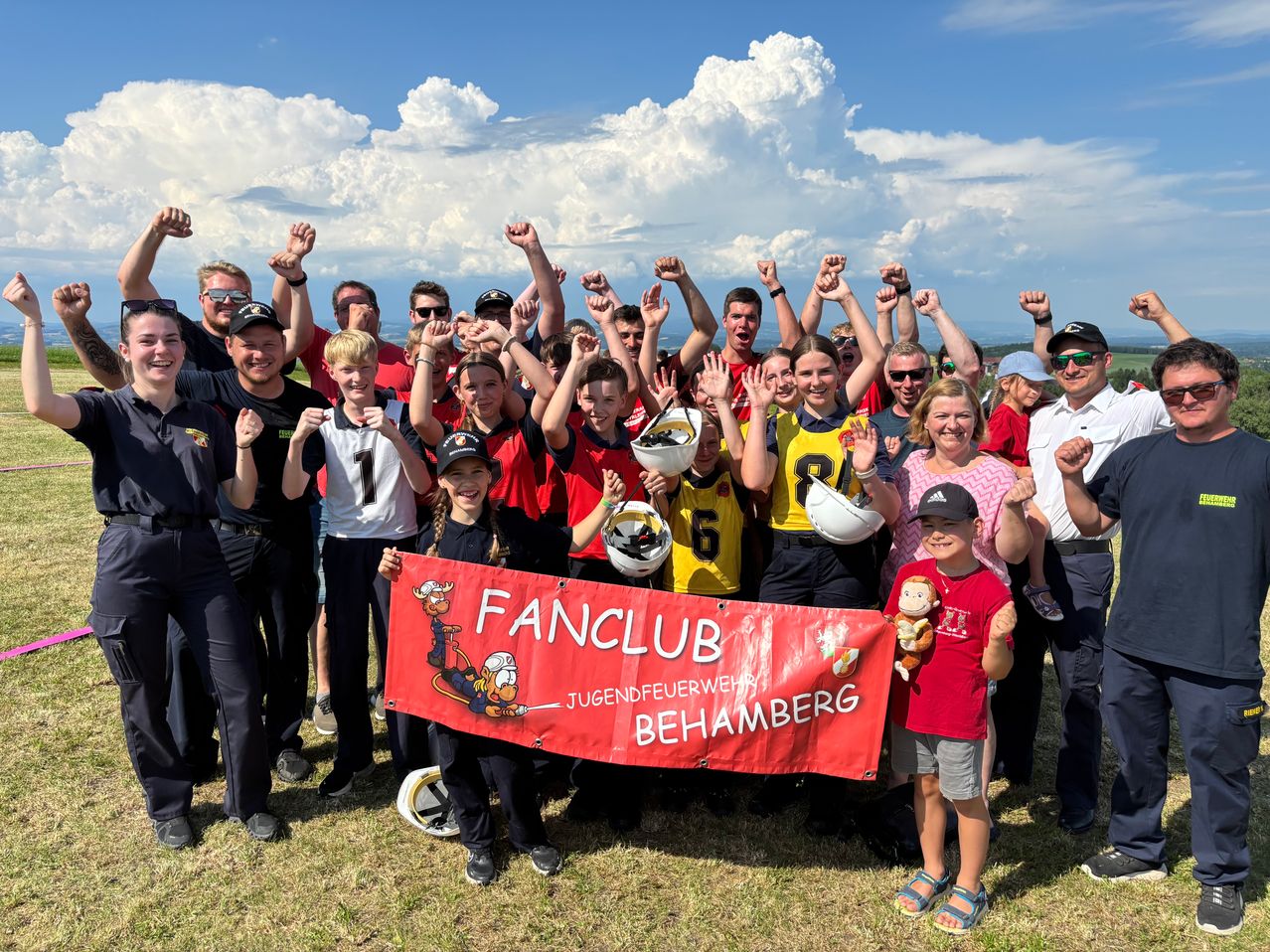 Eine Gruppe von Menschen mit Helmen und Trikots steht auf Gras und hält ein Banner, das Fanclub Jugendfeuerwehr Behamberg sagt. Sie posieren für ein Bild mit einem bewölkten Himmel.