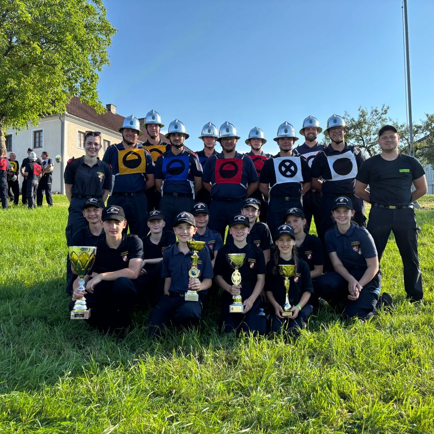 Eine Feuerwehrmannschaft posiert für ein Foto mit Pokalen, in Uniformen und Helmen, an einem sonnigen Tag.
