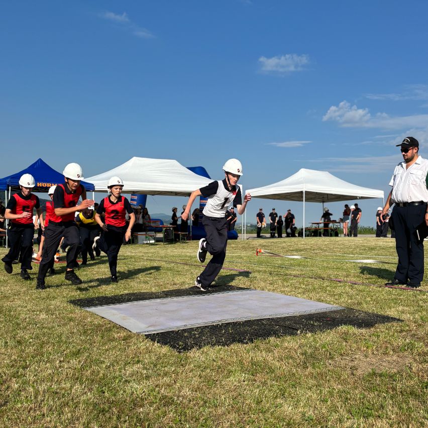 Eine Gruppe von Menschen in Helmen und roten und schwarzen Uniformen läuft auf einem Feld mit einem weißen Quadrat. Dahinter stehen Zelte und Menschen. Ein Mann in Hut und Uniform steht auf der rechten Seite.