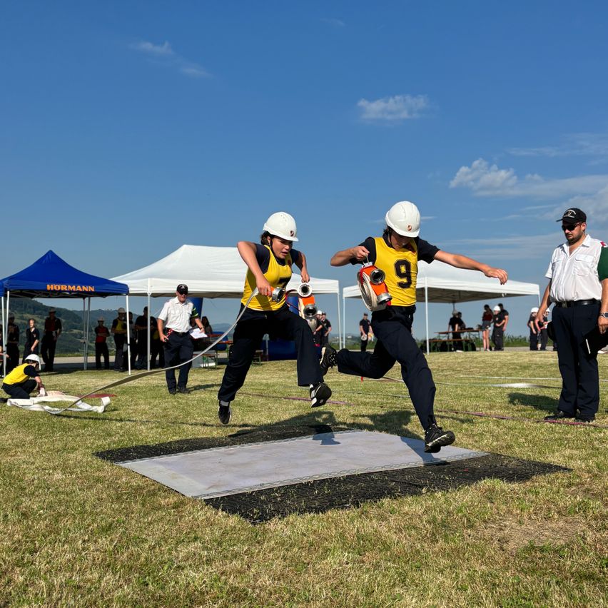 Zwei Personen in gelben Uniformen und Helmen führen einen Tauziehwettbewerb auf einem Feld durch. Zuschauer und Zelte im Hintergrund unter einem blauen Himmel.