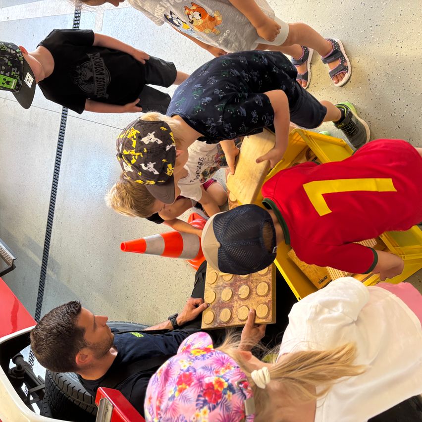Eine Gruppe von Kindern und ein erwachsener Mann versammeln sich um ein Holzbrett, möglicherweise in einer Werkstatt. Die Kinder schauen auf das Brett, während der Mann sitzt.