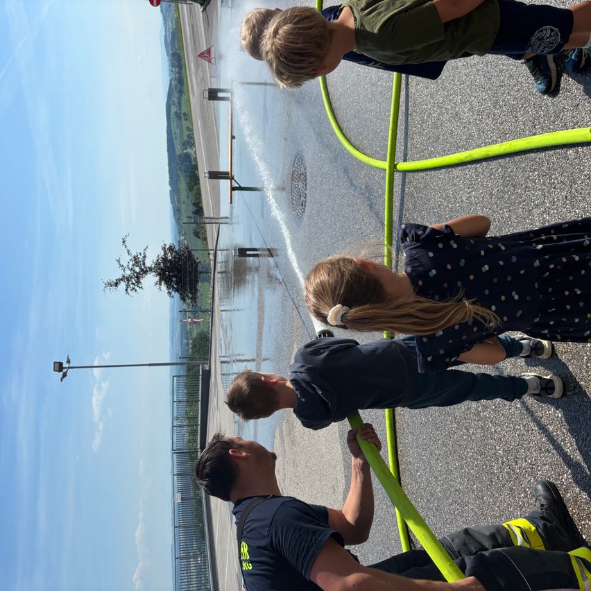 Drei Kinder und ein Feuerwehrmann spielen mit einem Schlauch, wobei ein Kind den Schlauch hält, während die anderen zuschauen. Die Szene spielt sich auf einer nassen Straße mit einem Zaun und Bäumen im Hintergrund ab.
