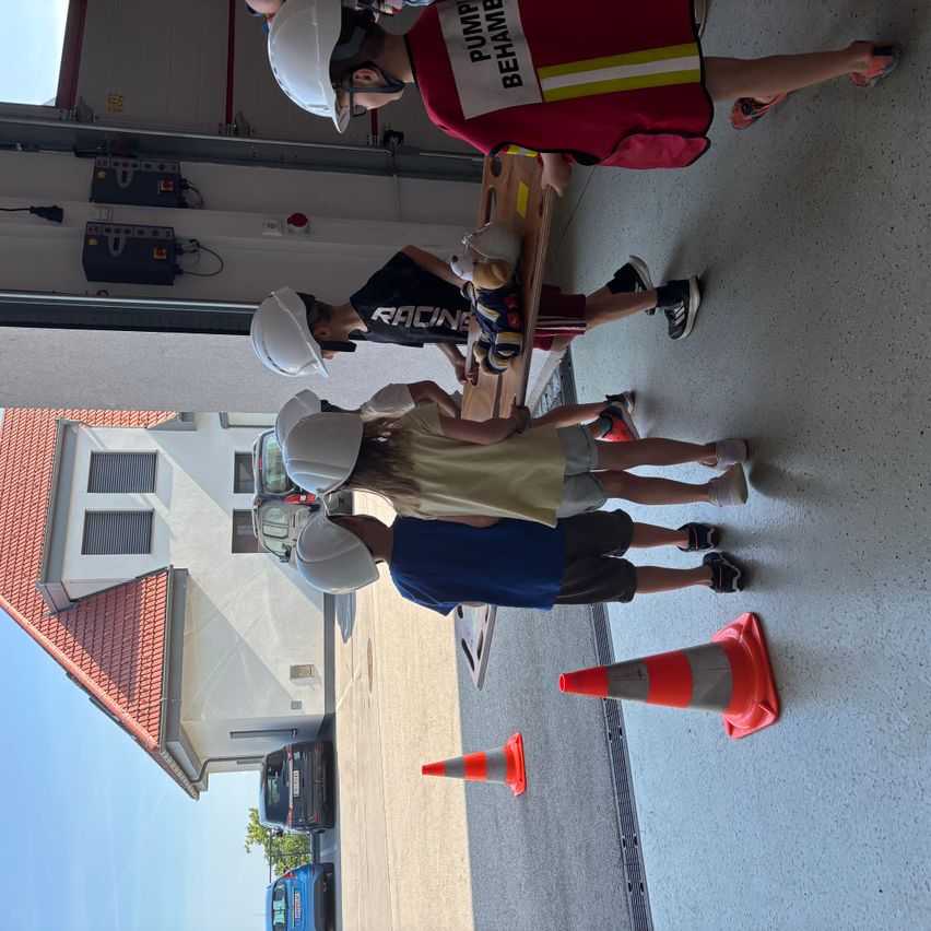 Fünf Kinder, einige in Helmen, üben Feuerwehrarbeit in einer Garage mit einem roten Haus dahinter.