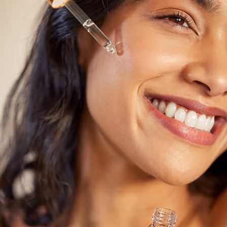 Eine Frau trägt mit einem Tropfenspender ein Serum auf ihr Gesicht auf und lächelt. Ihre Haut strahlt und ihre Zähne sind weiß.