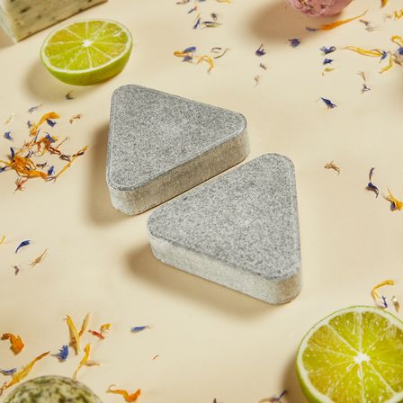 Zwei dreieckige Seifenstücke liegen auf einem Tisch, umgeben von Limettenscheiben und getrockneten Blütenblättern. Zusätzliche Seifenstücke sind im Hintergrund zu sehen.