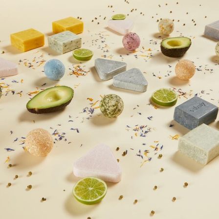 Verschiedene Seifen und Badeprodukte in unterschiedlichen Formen und Farben sind auf einer weißen Oberfläche verstreut. Avocadohälften, Limettenscheiben und Blumen sind ebenfalls vorhanden.