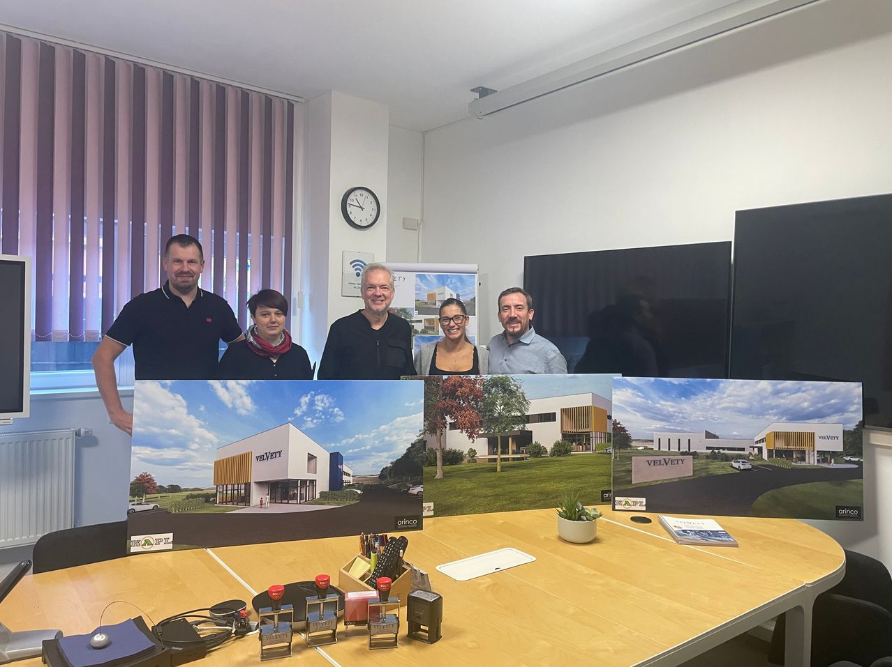 Five people stand behind a table displaying architectural plans, including a large image of a building, a book, and stamps. A woman wearing glasses stands in the center. A clock and a whiteboard hang on the wall.