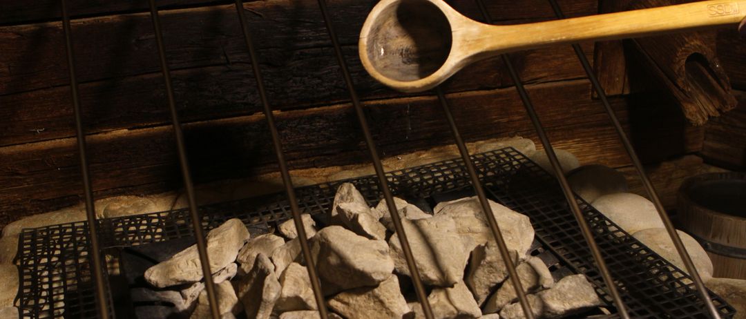 Ein hölzerner Löffel schwebt über einem mit Steinen gefüllten Grill, was auf die Vorbereitung eines Saunas hindeutet. Der Grill ist aus Metall mit einer hölzernen Wand im Hintergrund.