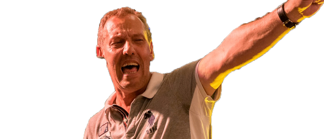 Ein Mann spielt auf einer Tastatur auf der Bühne und trägt ein Poloshirt mit Aufnähern. Er hat einen Arm erhoben und schreit.