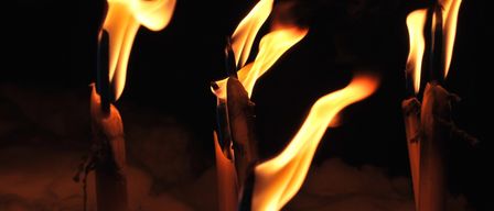 Drei brennende Kerzen leuchten hell, ihre Flammen ragen gegen einen dunklen Hintergrund nach oben. Die Kerzen haben ein rustikales Aussehen, mit ungleichmäßigen Flammen und einer leichten Rauchspur.