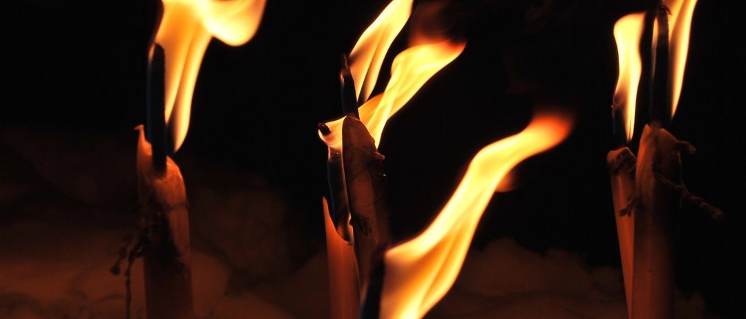 Drei brennende Kerzen leuchten hell, ihre Flammen ragen gegen einen dunklen Hintergrund nach oben. Die Kerzen haben ein rustikales Aussehen, mit ungleichmäßigen Flammen und einer leichten Rauchspur.