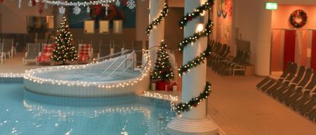 Ein Innenpoolbereich ist für Weihnachten dekoriert. Der Pool ist mit Girlanden geschmückt und es gibt Weihnachtsbäume mit Geschenken darum. Der Pool hat eine Spiralrutsche.