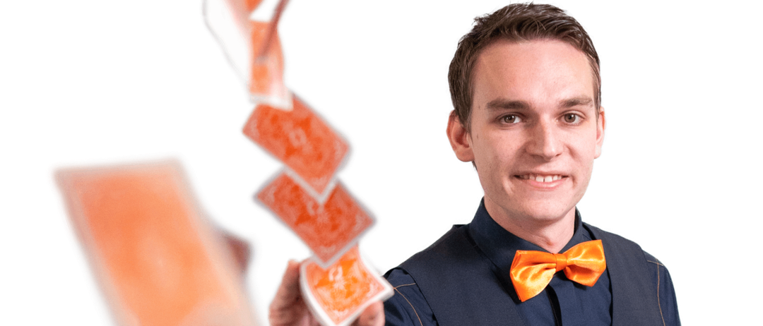 Ein Magier in einem blauen Anzug und oranger Fliege führt einen Kartentrick mit orangefarbenen Spielkarten in der Hand vor.