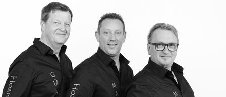 Drei Männer in schwarzen Jacken mit ihren Namen auf dem Rücken stehen zusammen und lächeln. Der Mann auf der linken Seite hat seine Hände in den Taschen. Der Mann in der Mitte hat seine Hände in den Taschen und schaut in die Kamera. Der Mann auf der rechten Seite trägt eine Brille und hat seine Hände in den Taschen.