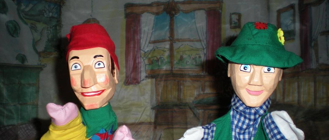 Zwei Puppen stehen nebeneinander vor einem bemalten Hintergrund. Die Puppe auf der linken Seite trägt einen roten Hut und ein gelb-blau gestreiftes Outfit. Die Puppe auf der rechten Seite trägt einen grünen Hut mit einer Blume und eine grüne Weste mit roten und gelben Aufnähern.