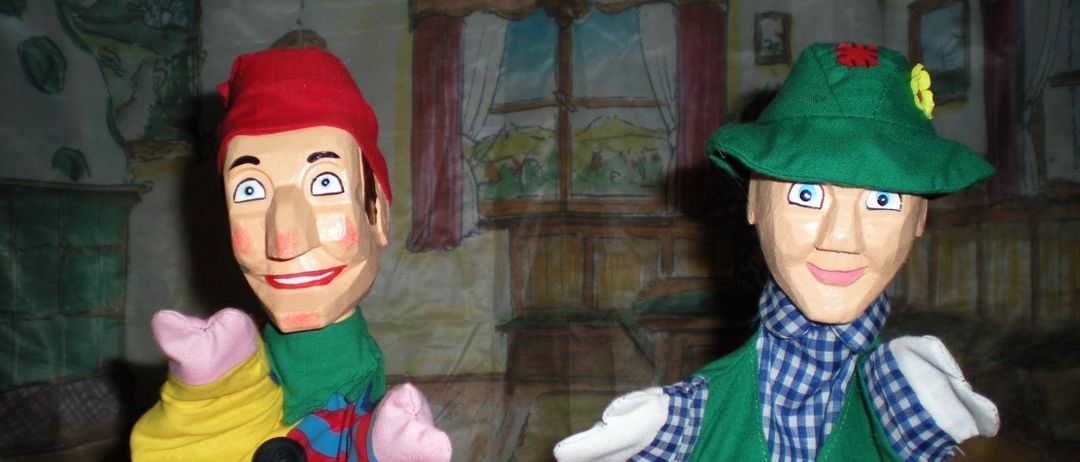 Zwei Handpuppen, eine mit rotem Hut und gelb-blau gestreiftem Shirt, die andere mit grünem Hut und grüner Weste, stehen vor einer Kulisse.