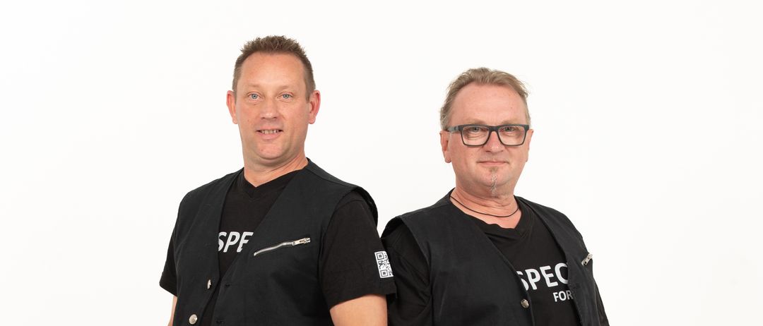 Zwei Männer stehen nebeneinander, beide tragen schwarze Westen mit weißem Text. Eine Weste sagt SPF und die andere SPEC. Sie tragen beide Jeans und haben die Hände in den Taschen.