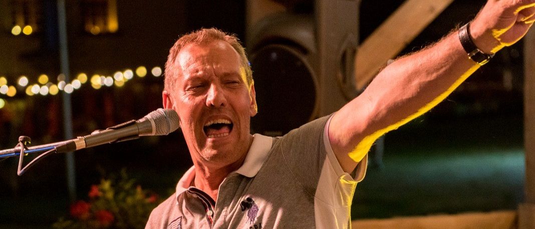 Ein Mann in einem grauen Polohemd mit Logo singt auf einer Bühne in ein Mikrofon. Er hat einen Arm erhoben und spielt eine Tastatur. Hinter ihm ist eine Holzwand und eine Leuchte auf dem Boden.