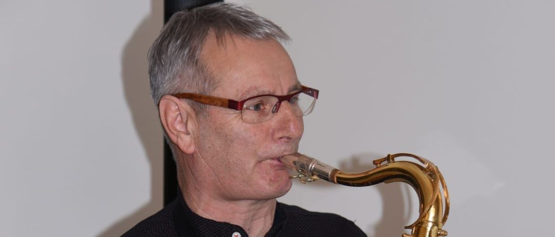 Ein älterer Mann mit Brille spielt eine goldene Saxophon. Er trägt ein schwarzes Button-Down-Hemd und einen schwarzen Riemen über der Schulter. Der Hintergrund ist weiß.