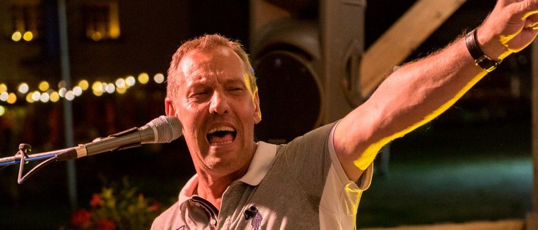 Ein Mann singt in ein Mikrofon auf der Bühne mit einem Klavier vor ihm. Er hat eine Uhr und ein Armband an.