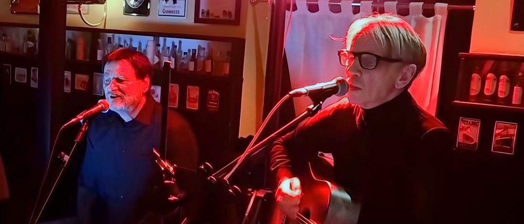 Zwei Männer treten in einer Kneipe mit roter Beleuchtung auf. Einer spielt Gitarre und singt in ein Mikrofon, während der andere neben ihm sitzt. Hinter ihnen befinden sich Regale mit Flaschen, gerahmte Bilder und ein Schild für '100 Whiskeys'.