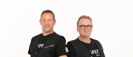 Zwei Männer stehen nebeneinander in identischen schwarzen Westen mit weißem Text. Sie stehen vor einem schlichten weißen Hintergrund.