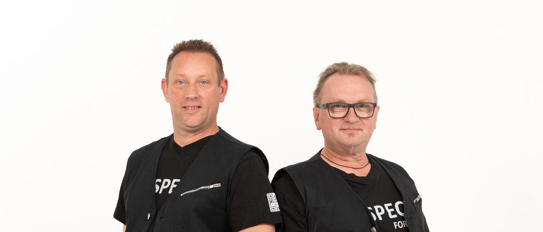 Zwei Männer stehen nebeneinander in identischen schwarzen Westen mit weißem Text. Sie stehen vor einem schlichten weißen Hintergrund.