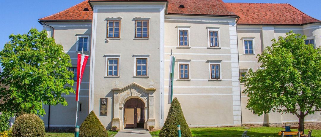 Ein großes historisches Gebäude mit rotem Ziegeldach und weißen Wänden. Es hat viele Fenster und zwei Fahnen auf Stangen. Ein Weg führt zum gewölbten Eingang.