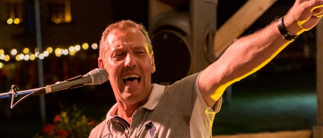 Ein Mann singt in ein Mikrofon auf einer Bühne. Er trägt ein graues Shirt mit weißem Saum.