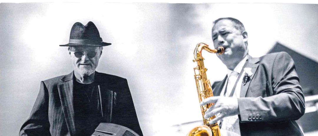 Zwei Musiker, einer mit einem Saxophon und der andere mit einer Tastatur, stehen draußen. Der Mann mit dem Saxophon konzentriert sich auf sein Instrument, während der andere eine Tastatur hält.