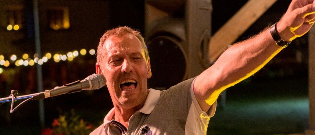 Ein Mann singt in ein Mikrofon, steht vor einem Klavier, mit einem erhobenen Arm und einem Holzzaun dahinter.