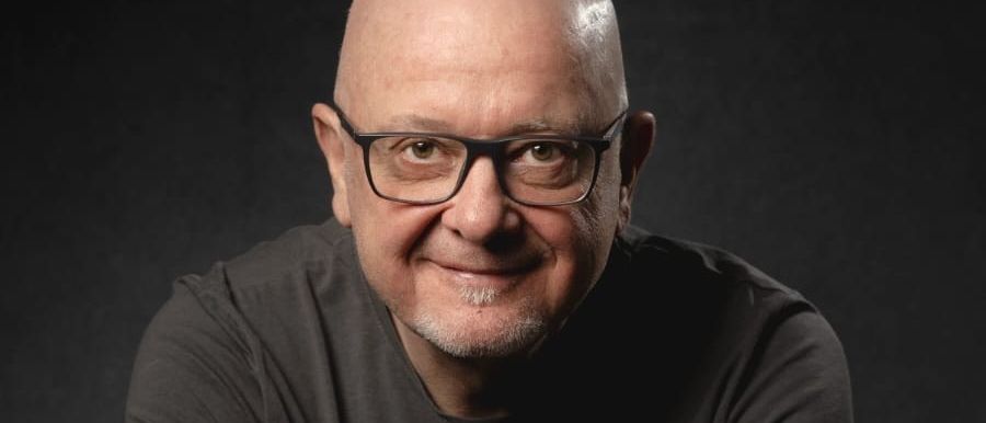 Ein lächelnder Glatzkopf mit Brille und schwarzem Shirt posiert mit verschränkten Händen vor dunklem Hintergrund.
